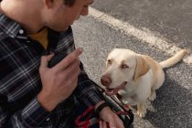 Високий кут зору крупним планом молодого Кавказького людини в інвалідному візку дивлячись на його собаку, сидячи на тротуарі дивлячись на нього — стокове фото