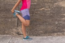 Вид сбоку низкая секция женщины в спортивной одежде, стоящей перед стеной на улице, держа ногу и растягивая ногу во время тренировки — стоковое фото
