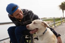 Вид спереду крупним планом молодого Кавказького чоловіка в інвалідному візку приймаючи прогулянку зі своєю собакою в сільській місцевості, посміхаючись — стокове фото