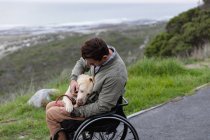 Side view of a young Caucasian man in a wheelchair taking a walk with his dog in the countryside by the sea, stroking the dog on his lap — Stock Photo