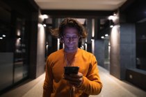 Vista frontal de perto de um jovem caucasiano na rua à noite olhando para um smartphone e usando fones de ouvido — Fotografia de Stock