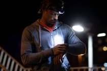 Vista frontal close-up de um jovem caucasiano verificando um smartwatch na rua durante seu treino noturno com um farol ligado — Fotografia de Stock