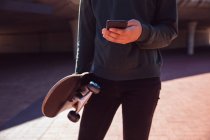 Vorderansicht Mittelteil eines modischen Mannes auf der Straße, der ein Smartphone benutzt und ein Skateboard in der Hand hält — Stockfoto