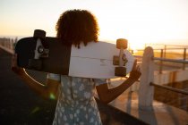 Vista posteriore da vicino della giovane donna di razza mista che tiene uno skateboard sulle spalle ammirando la vista sul mare, retroilluminata dal sole al tramonto — Foto stock