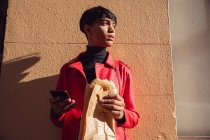 Vista lateral de um jovem elegante mestiço adulto transexual na rua, usando um smartphone e comendo um sanduíche — Fotografia de Stock
