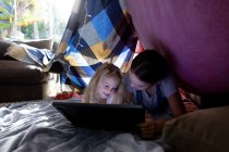 Vista frontal de una joven chica caucásica y su hermana adolescente usando una tableta juntos en una tienda hecha de mantas - foto de stock
