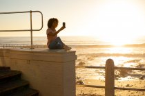 Vista lateral da jovem mulher mista tirando uma selfie, sentada em uma parede ao pôr do sol junto ao mar — Fotografia de Stock