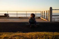 Front view of a pre-teen boy sitting on a skateboard at sunset by the sea — Stock Photo