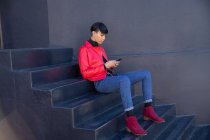 Vista lateral de um jovem adulto transexual de raça mista na moda na rua, sentado em passos usando um smartphone contra uma parede cinza — Fotografia de Stock