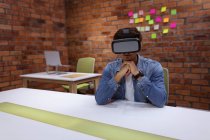 Front view of a young Caucasian man sitting at a desk wearing a VR headset with his chin resting on his hands, in the office of a creative business — Stock Photo