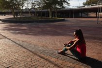 Vista lateral de uma jovem caucasiana vestindo roupas esportivas sentada no chão verificando seu smartwatch enquanto trabalhava em um dia ensolarado em um parque — Fotografia de Stock