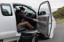 Вид сбоку на молодого кавказца, выходящего из инвалидного кресла и садящегося в машину на стоянке у моря — стоковое фото