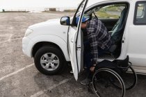 Vista lateral de un joven caucásico bajándose de un coche en una silla de ruedas en un aparcamiento junto al mar - foto de stock