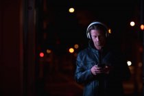 Vorderansicht eines jungen kaukasischen Mannes, der abends auf einer Straße steht und mit aufgesetzten Kopfhörern Musik hört und ein Smartphone in der Hand hält — Stockfoto