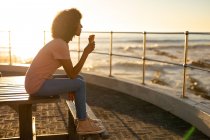 Вид збоку на молоду змішану жінку, яка сидить на лавці, їсть морозиво і захоплюється видом на захід сонця біля моря — стокове фото