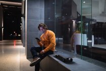 Vista lateral de um jovem caucasiano sentado em uma parede por um prédio à noite com fones de ouvido em ouvir música e olhar para um smartphone, com um skate ao lado dele — Fotografia de Stock