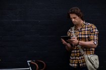 Vista frontal de un joven caucásico de pie en la calle sosteniendo una tableta con una bicicleta a su lado durante su viaje a casa por la noche - foto de stock