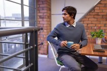 Фронт-вью крупным планом молодого кавказца, сидящего за столом, пьющего кофе и поворачивающегося, чтобы посмотреть в окно в офисе творческого бизнеса — стоковое фото