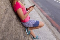 Вид збоку низька частина жінки в спортивному одязі, що спирається на стіну на вулиці, перевіряючи її смартфон під час роботи — стокове фото
