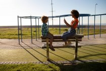 Vista lateral de una joven mestiza y su hijo preadolescente disfrutando del tiempo juntos jugando en un parque infantil junto al mar, sentados en un banco y tomando fotos en un día soleado con un marco de escalada en el fondo - foto de stock