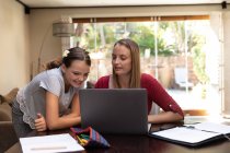 Front view of a young Caucasian woman using a laptop computer with her tween daughter looking on, in their dining room — Stock Photo