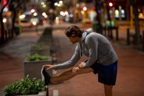 Seitenansicht eines jungen kaukasischen Mannes, der sich während seines späten Abendtrainings auf der Straße dehnt — Stockfoto