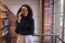 Vista frontal close-up de uma jovem mulher de raça mista sorridente de pé e falando em um smartphone no escritório de um negócio criativo — Fotografia de Stock