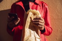 Вид спереди среднего сегмента модного мужчины на улице, с помощью смартфона и едят сэндвич — стоковое фото
