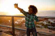 Вид спереду усміхненого хлопчика, який тримає смартфон і тримає селфі на балюстраді на заході сонця біля моря — стокове фото