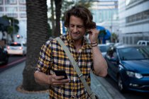 Вид спереду крупним планом молодого Кавказького чоловіка тримає смартфон і поклавши навушники на жвавій міській вулиці ввечері — стокове фото