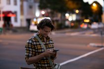 Vista lateral de un joven hombre caucásico enviando mensajes de texto de pie en una calle de la ciudad durante su viaje nocturno - foto de stock