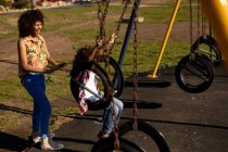 Vista lateral de una joven mestiza y su hijo preadolescente disfrutando del tiempo juntos jugando en un parque infantil, el niño balanceándose en un día soleado - foto de stock