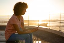 Seitenansicht einer lächelnden jungen Mischlingshündin mit einem Smartphone, die bei Sonnenuntergang am Meer auf einer Bank sitzt — Stockfoto