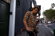 Nahaufnahme eines jungen kaukasischen Mannes, der sich auf ein Fahrrad stützt und abends ein Smartphone in einer Straße in der Stadt benutzt — Stockfoto