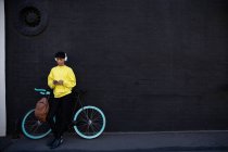 Передній вигляд модного молодого змішаної раси транссексуал дорослий на вулиці, смс на смартфоні, що стоїть поруч з велосипедом з навушниками на сірій стіні — стокове фото