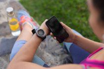 Par-dessus l'épaule de la femme portant des vêtements de sport vérifiant sa montre intelligente et son smartphone tout en s'entraînant dans un parc — Photo de stock