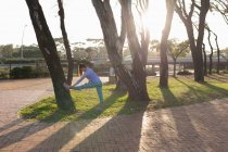 Вид збоку молоді кавказька жінка носіння спортивного одягу спираючись на дерево розтягуючи ногу під час тренування в парку — стокове фото