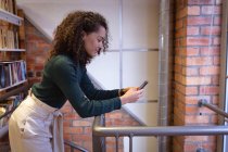 Зворотний вид усміхнена молода змішана раса жінка стоїть і використовує смартфон в офісі творчого бізнесу — стокове фото