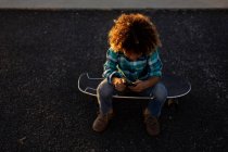 Hochwinkelaufnahme eines Jungen im Teenageralter, der mit einem Smartphone auf einem Skateboard sitzt — Stockfoto