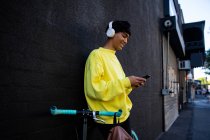 Vista lateral de um jovem elegante mestiço transexual adulto na rua, mensagens de texto no smartphone ao lado de uma bicicleta com fones de ouvido em — Fotografia de Stock