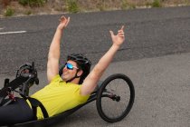 Вид сбоку на молодого кавказца в спортивной одежде на лежачем велосипеде, катающегося на велосипеде по проселочной дороге, улыбающегося с поднятыми руками — стоковое фото