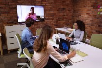 Сторона зору молодої змішаної раси і молодої кавказької жінки і чоловіка, які працюють на ноутбуках в офісі творчого бізнесу. Чоловічий колега видно на екрані, встановленому на стіні, спілкуючись за допомогою відеозв 