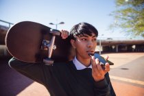Модный молодой трансгендер смешанной расы на улице, разговаривающий по смартфону и держащий в руках скейтборд — стоковое фото