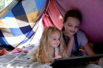 Vue de face d'une jeune fille caucasienne et sa soeur entre deux en utilisant une tablette ensemble dans une tente faite de couvertures — Photo de stock