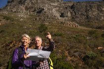 Vista frontale da vicino di un uomo e una donna caucasici maturi che leggono una mappa e puntano durante una passeggiata in un ambiente rurale, con le montagne sullo sfondo — Foto stock
