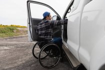 Vista lateral de un joven caucásico en silla de ruedas junto a su coche, estacionado en un aparcamiento con la puerta abierta - foto de stock
