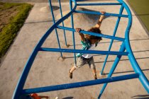 Vista de ángulo alto de un niño preadolescente de raza mixta jugando en un patio de recreo, colgado de un marco de escalada en un día soleado - foto de stock