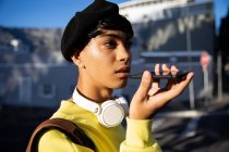 Вид сбоку на модную молодую смешанную расу трансгендерного взрослого на улице, разговаривающего по смартфону в берете — стоковое фото