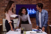 Вид спереди на улыбающуюся молодую женщину смешанной расы и молодую кавказскую женщину и мужчину, стоящих рядом с ноутбуком, работающих вместе в офисе творческого бизнеса, смотрящих друг на друга и улыбающихся — стоковое фото