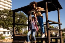 Front view of a young mixed race woman and her pre-teen son enjoying time together playing at a playground, standing on a climbing frame on a sunny day with trees and buildings in the background — Stock Photo